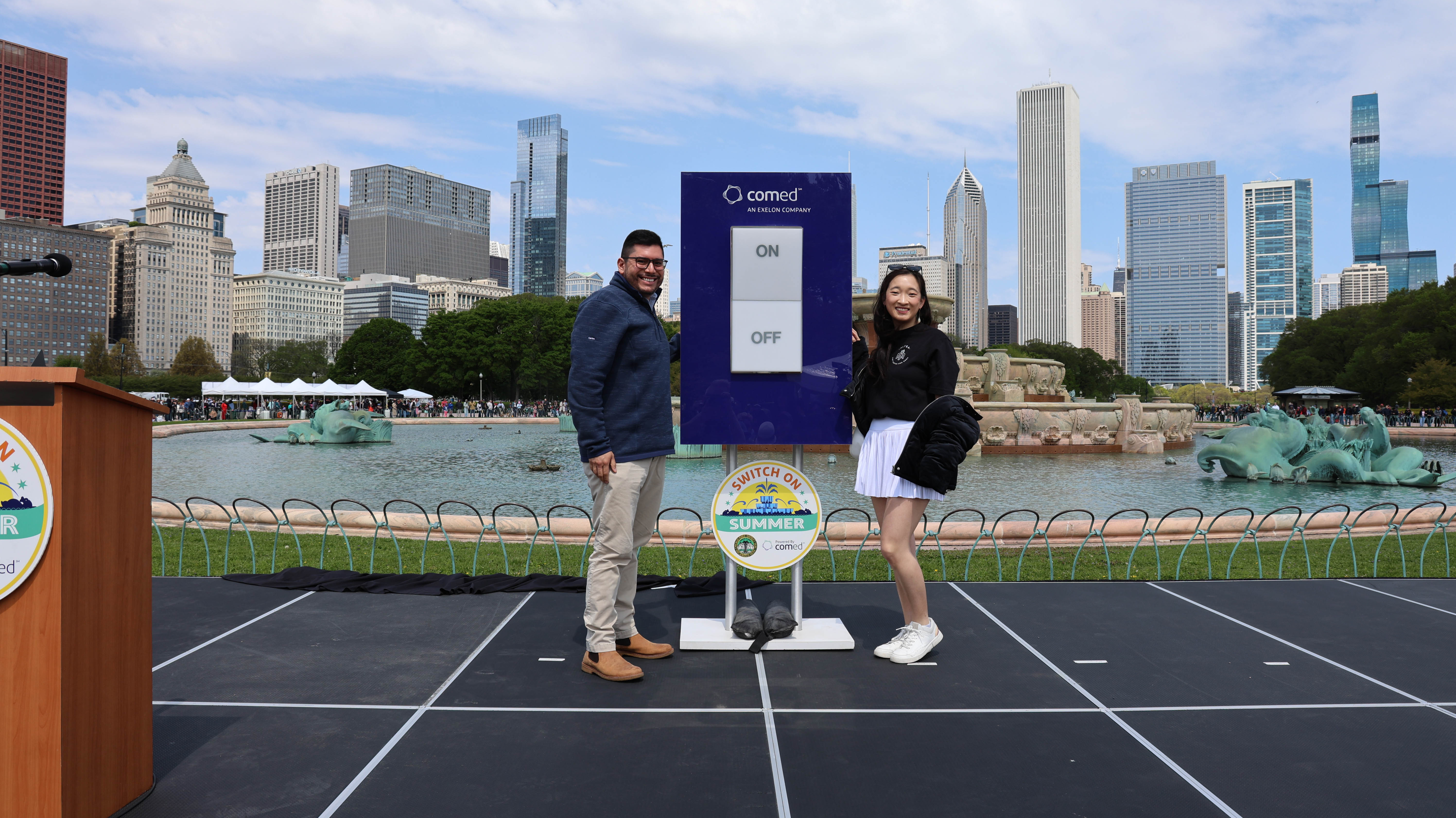 Two people stand on a stage by Buckingham Fountain in Chicago with a large ComEd switch.
